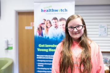 Young girl stood in a pink hoodie in front of a Healthwatch banner Young girl stood in a pink hoodie in front of a Healthwatch banner
